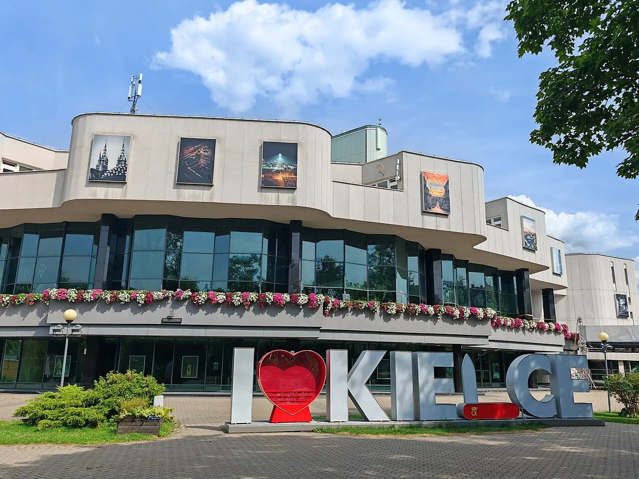 A modern building with a curved facade and large windows, adorned with flower boxes, stands behind large letters on the ground spelling out "I ❤️ KIELCE."
