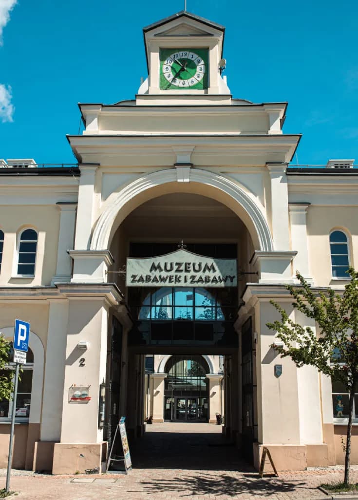 A cream-colored building with a large arched entrance and a clock tower above, under a clear blue sky, with a sign that reads "MUZEUM ZABAWEK I ZABAW".