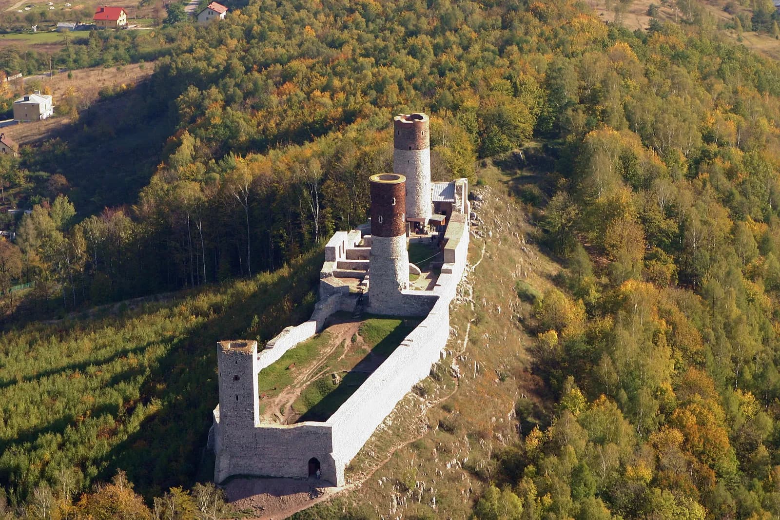 An aerial view of the ruins of a stone castle with two prominent towers situated on a forested hill, with some houses visible in the distance.